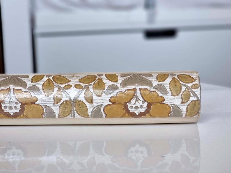 May include: A cylindrical rolling pin featuring a repeating floral pattern with mustard yellow, brown, and muted green leaves and petals against a white background. The surface has a textured, wood-grain appearance with carved details.