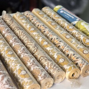 May include: A collection of nine rolls of wallpaper arranged in a row. Each roll features distinct patterns in shades of mustard yellow, brown, beige, and white, including floral, damask, and geometric motifs. One roll has a blue and green label.