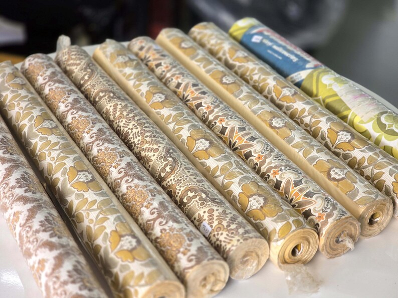 May include: A collection of nine rolls of wallpaper arranged in a row. Each roll features distinct patterns in shades of mustard yellow, brown, beige, and white, including floral, damask, and geometric motifs. One roll has a blue and green label.