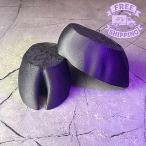 May include: Two dark gray, rock-shaped objects are displayed on a textured, gray surface. The objects have a smooth, rounded top and a split, curved bottom. The lighting creates a gradient effect, with the objects appearing darker at the base and lighter at the top.