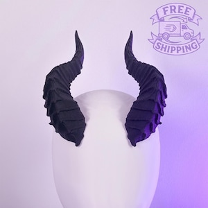 Könnte beinhalten: Ein Paar schwarze, strukturierte Hörner, wahrscheinlich für ein Kostüm oder Cosplay, werden auf einer weißen, abgerundeten Form präsentiert. Die Hörner krümmen sich nach oben und außen, mit einer geriffelten Textur. Der Hintergrund ist ein Farbverlauf aus Weiß und Lila.