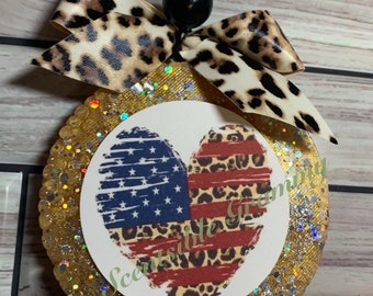 May include: A round, gold glitter car air freshener with a white heart-shaped design. The heart is divided in half, with one side featuring an American flag design and the other side featuring a leopard print design. The air freshener is tied with a leopard print ribbon.