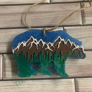 May include: A blue, green, brown, and white bear ornament with a mountain and tree design. The ornament is hanging from a twine string.