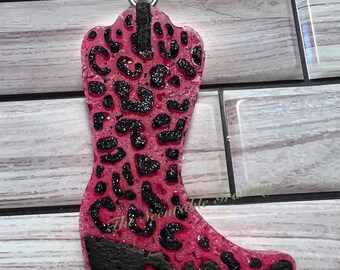 May include: Pink cowboy boot shaped car air freshener with a black leopard print design. The boot is hanging from a black string.