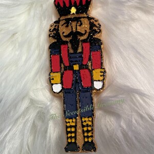 May include: A decorative nutcracker figurine with a gold outline, red and blue uniform, and a black mustache. The figurine has a red crown and is on a white, fluffy surface. The text "The Scentsible Granny" is visible.