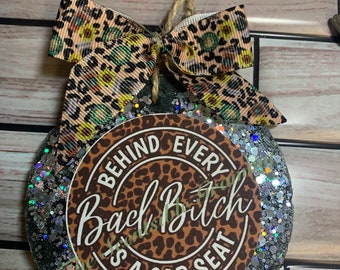May include: A round, black, glitter-covered ornament with a brown circle in the center. The brown circle has a leopard print pattern and the text "Behind Every Bad Bitch Is A Car Seat". The ornament is decorated with a brown and black leopard print ribbon.