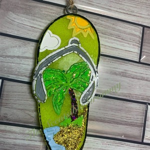 May include: A green flip-flop shaped ornament with a palm tree, a sun, and a cloud. The ornament is decorated with glitter and has a string for hanging.