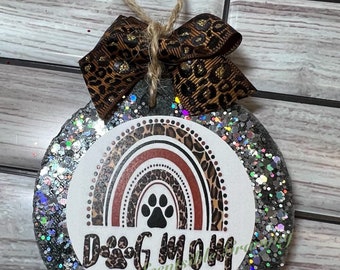 May include: A round, silver glitter ornament with a white circle in the center. The circle features a brown, orange, and white rainbow with a black paw print in the center. The text "Dog Mom" is written in black with a paw print replacing the letter "o". The ornament is tied with a brown twine and a brown and gold leopard print ribbon.
