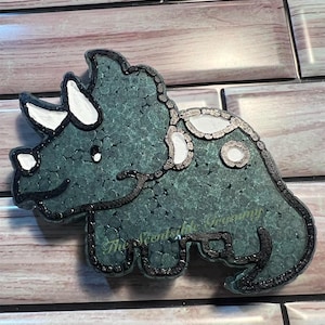 May include: A green and black three-horned dinosaur figurine with white spots. The dinosaur is made of a textured material and has a cartoonish style.