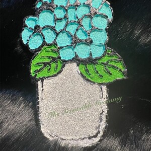 May include: A decorative pin featuring a turquoise hydrangea flower with black outlines and green leaves, set in a silver-colored vase. The text "The Wearable Mommy" is printed on the vase.