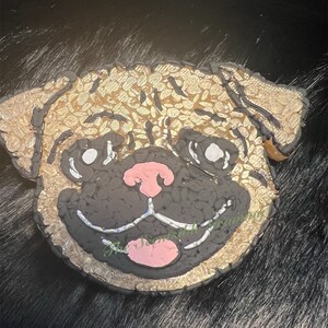 May include: A close-up of a pug dog face made of mosaic tiles. The tiles are gold, black, and white. The dog's nose is pink.