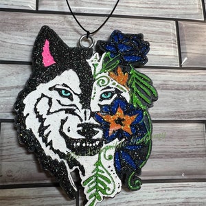 May include: A black and white wolf with glitter, with a blue and green floral design on the right side of its face. The wolf is a car air freshener with a black string.