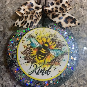 May include: Round, glitter-covered car air freshener with a sunflower and bee design. The center features a yellow sunflower with a bee, the text "Be Kind", and a leopard print bow.
