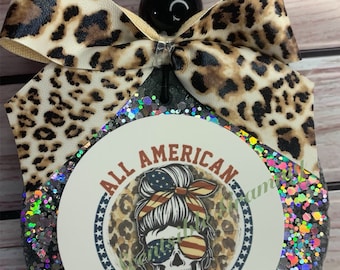 May include: A round, black, glitter-covered ornament with a white circle in the center. The white circle has a graphic of a woman with a skull face wearing a red, white, and blue bandana and sunglasses. The text "All American Mama" is printed around the graphic.