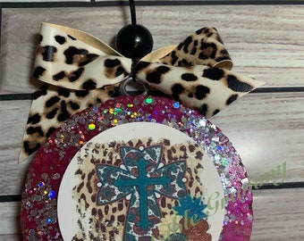 May include: A round, pink glittered car air freshener with a leopard print bow and a black string. The center features a white circle with a turquoise cross design and floral accents. The text "The Little Granny" is visible.