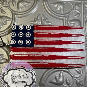 May include: A red, white, and blue American flag with a distressed look. The flag is made of wood and has a white background with red stripes and blue stars. The stars are arranged in a circle in the upper left corner of the flag. The flag is hanging from a string. The text "The Scentsible Grammy" is on a white circle with a black and white leopard print background.