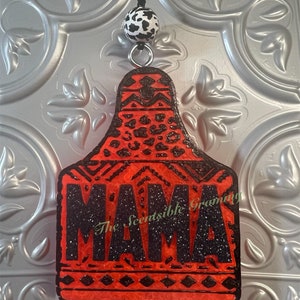 May include: Red and black car air freshener with a cow print bead hanging from it. The air freshener is shaped like a cattle tag and says "MAMA" in black glitter. The text "The Scentsible Granny" is also visible.