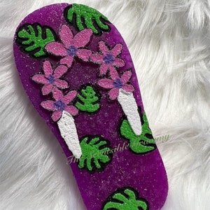 May include: Purple glitter flip flop shaped soap with green and black leaves and pink flowers. The soap has the text "The Scentsable Granny" on it.