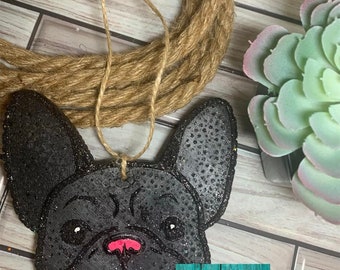 May include: Black glitter French bulldog car air freshener with a pink nose and white eyes. The air freshener is hanging from a twine string. The text "The Scentsible Grammy" is on a small tag in the bottom right corner of the image.