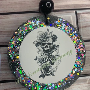 May include: A round black car air freshener with a white circle in the center. The white circle has a black and white image of a skull with roses. The outer edge of the air freshener is covered in iridescent glitter.