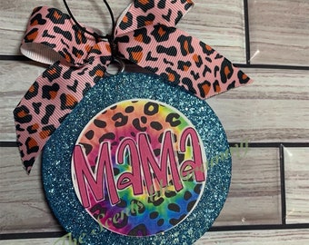 May include: A teal glitter round ornament with a pink, blue, and orange tie-dye design featuring a black leopard print. The word "MAMA" is written in pink letters on the ornament. The ornament is tied with a pink and black leopard print ribbon.