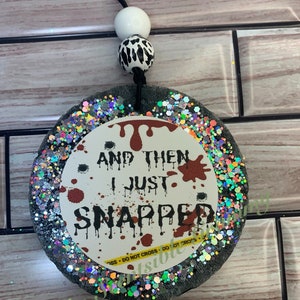 May include: A round black and silver glitter car air freshener with a white circle in the center. The white circle has red blood splatter and the text "And then I just snapped".