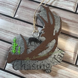 May include: A silver and brown ornament with a deer antler design and the text "The Scentsable Garden Chasing Tail" in black lettering.