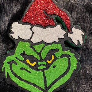 May include: A Grinch-themed ornament featuring a green face with yellow eyes and a red glittery Santa hat. The ornament has a black outline and a silver loop for hanging. The Grinch's face is detailed with glitter.