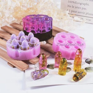 Puede incluir: Una colección de moldes de resina y piezas de resina terminadas. Los moldes son rosas y morados, y las piezas terminadas tienen forma de bala con detalles dorados. El texto "Daisy Note Craft" es visible.
