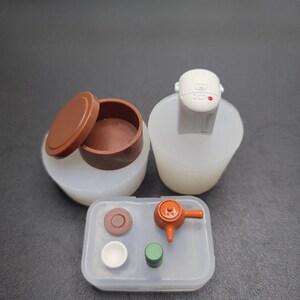 以下が含まれることがあります： 炊飯器、蓋付き容器、ティーポット、カップ、その他の小さなアイテムが入ったトレイなど、ミニチュアのおもちゃのキッチンアイテムのコレクション。アイテムは茶色、白、オレンジの色合いです。