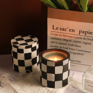 May include: Two candles with a black and white chequered pattern. One is lit, the other has a lid. The background includes a paper bag with French text.