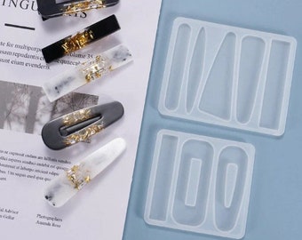 Könnte beinhalten: Eine Auswahl handgefertigter Haarspangen in Schwarz, Weiß und Grau, verziert mit Blattgold. Zwei Silikonformen zur Herstellung von Haaraccessoires sind ebenfalls zu sehen, mit DIY-Thema.