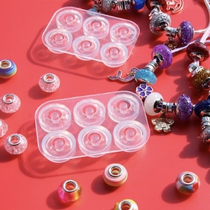 May include: A collection of colorful charm bracelets and individual beads on a red surface. The bracelets feature various charms, including glittery beads, flowers, and sea creatures. Clear plastic molds with circular indentations are also present.
