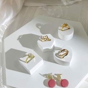 Puede incluir: Una colección de anillos dorados y pendientes rosas exhibidos en soportes geométricos blancos. Los anillos presentan varios diseños, incluidos motivos florales y de corona. Los pendientes tienen una gota circular rosa con una parte superior dorada.