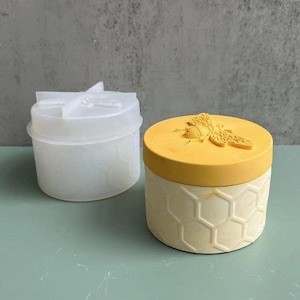 Puede incluir: Un recipiente redondo amarillo y crema con un diseño de abeja en la tapa. La tapa es amarilla y el cuerpo del recipiente tiene un patrón de panal. Un molde de plástico transparente está al lado del recipiente.