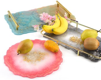 May include: Decorative resin trays in shades of blue, gray, and pink, with gold-colored handles. The trays are adorned with gold flakes and hold fresh fruit, including bananas, pears, and kiwi. The trays have a unique, organic shape.