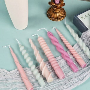 May include: A collection of pastel-colored spiral and twisted candles. The candles vary in shades of pink, green, and peach. A gold candle holder and a white vase are also visible. The candles are arranged on a light blue surface with a white lace runner.