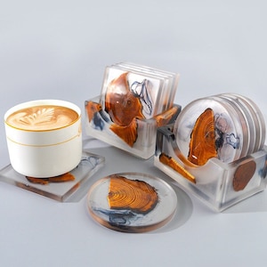 Può includere: Un set di sottobicchieri con finitura in resina trasparente, che mostra fette di legno naturale e un effetto marmorizzato. Il set include sottobicchieri quadrati e rotondi, con una tazza di caffè sopra. I sottobicchieri sono esposti in supporti trasparenti.