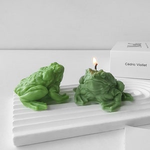 Könnte beinhalten: Zwei grüne, froschförmige Kerzen auf einem weißen, geriffelten Tablett. Eine Kerze ist angezündet, mit einer kleinen Flamme. Die Kerzen haben eine strukturierte Oberfläche und stehen neben einer weißen Box mit dem Text "Cédric Viollet".