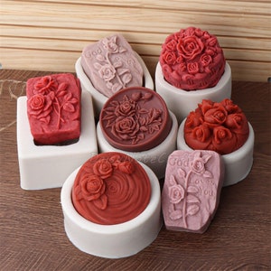 May include: A selection of rose-shaped soaps in shades of red and pink, presented in white moulds. Some soaps feature the text "God Loves You" and "Loves You". The soaps are in rectangular, round, and dome shapes.