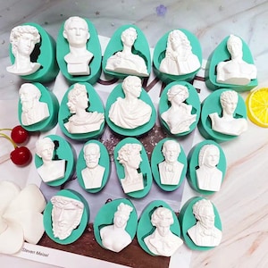Può includere: Una collezione di stampi per busti in gesso bianco in vari stili classici, inseriti in stampi in silicone ovali color verde acqua. I busti raffigurano figure storiche e sono disposti su una superficie a motivi.