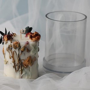May include: A white candle decorated with dried flowers and greenery, next to a clear cylindrical container. The candle has a wick and is adorned with various dried floral elements in shades of brown, red, and green.