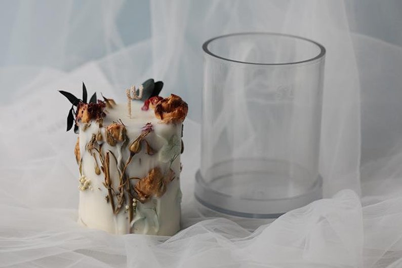 May include: A white candle decorated with dried flowers and greenery, next to a clear cylindrical container. The candle has a wick and is adorned with various dried floral elements in shades of brown, red, and green.