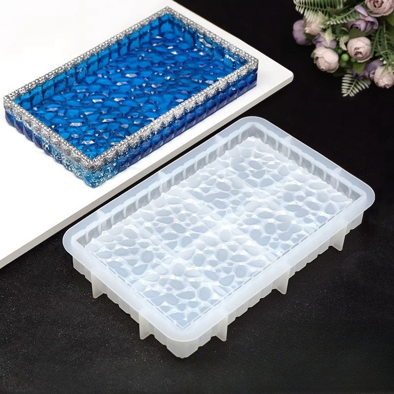 May include: A rectangular resin tray with a blue, gem-like interior and a silver glitter border. A clear silicone mold with a pebble-like pattern is in the foreground. The tray is on a white surface.