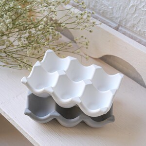 May include: Two-tone egg holder in white and grey. The top tier is white, and the bottom tier is grey. The holder has a wavy design with six egg compartments. The background includes white flowers and a wooden surface.