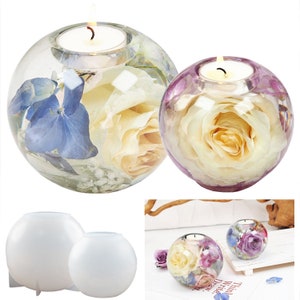 May include: Two spherical candle holders with dried flowers encased in clear resin. One has white roses and blue hydrangeas, the other has a white rose. A lit candle sits inside each. Two white spherical molds are also shown.