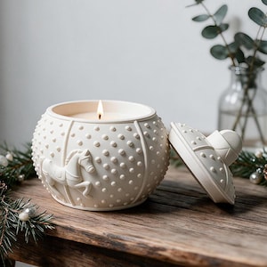 May include: A round, off-white ceramic candle jar featuring a textured surface with raised dots and a relief of a carousel horse. The jar is lit with a single flame and includes a matching removable lid resting beside it on a wooden surface.
