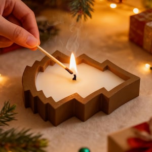 Könnte beinhalten: Eine brennende Kerze in Form eines Weihnachtsbaums mit braunem Rahmen. Ein Streichholz wird in die Nähe des Dochts gehalten, Rauch steigt auf. Die Kerze steht auf einer weißen Oberfläche, mit Weihnachtsdekorationen im Hintergrund.