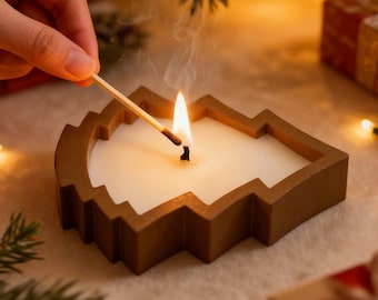 May include: A lit Christmas tree-shaped candle with a brown frame. A match is held near the wick, with smoke rising. The candle is on a white surface, with Christmas decorations in the background.