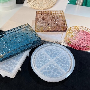 May include: A collection of resin molds and finished resin pieces. The molds are white and the finished pieces are blue, pink, and brown with gold accents. The pieces have a textured surface and are square and round.